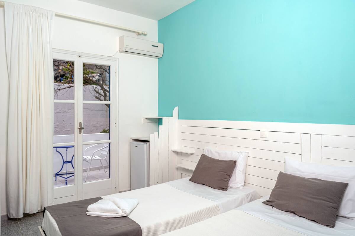 A room with two beds, a window with curtains, a small table, and an air conditioning unit.