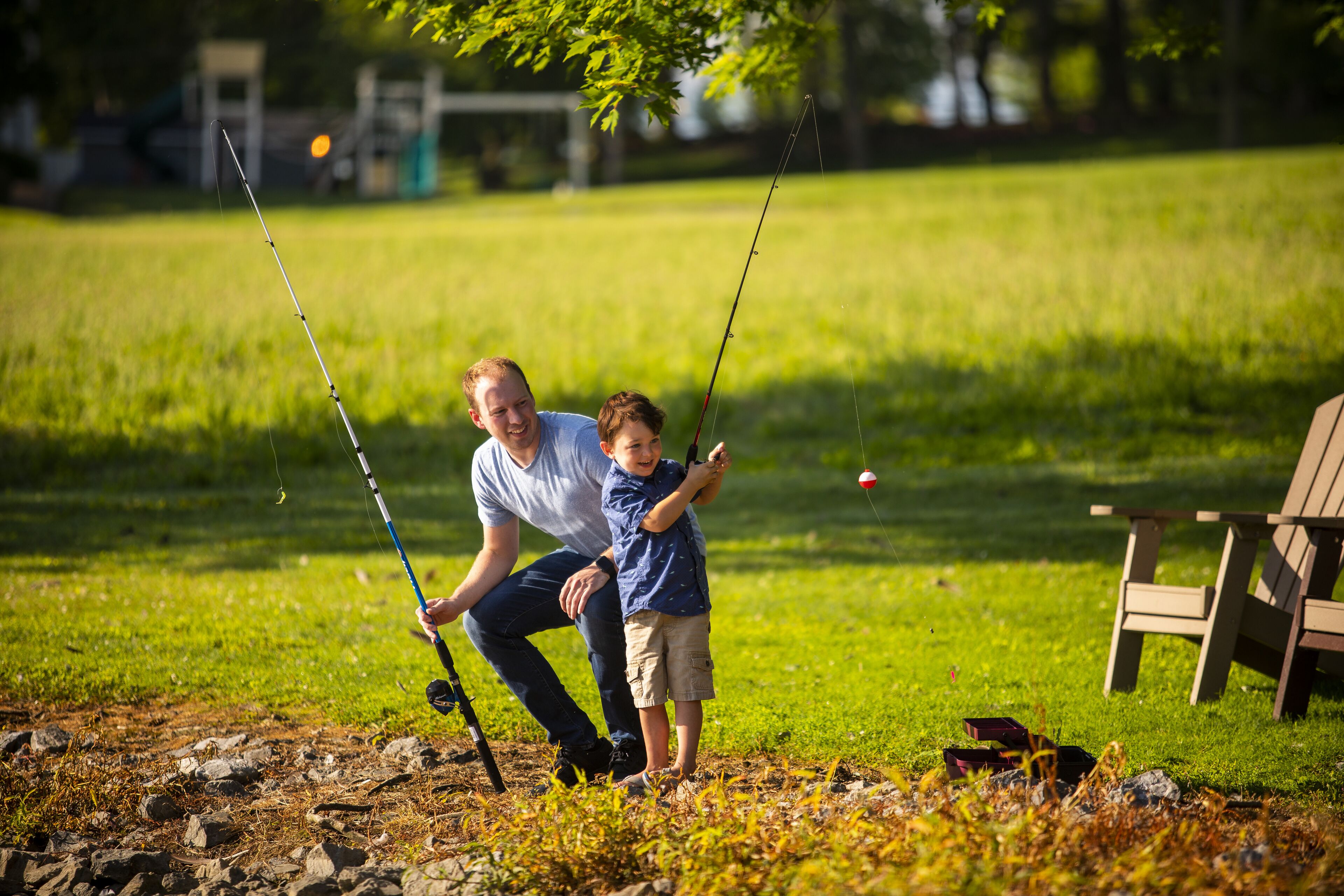 A man and a child fishing in a grassy area with a picnic table in the background.
