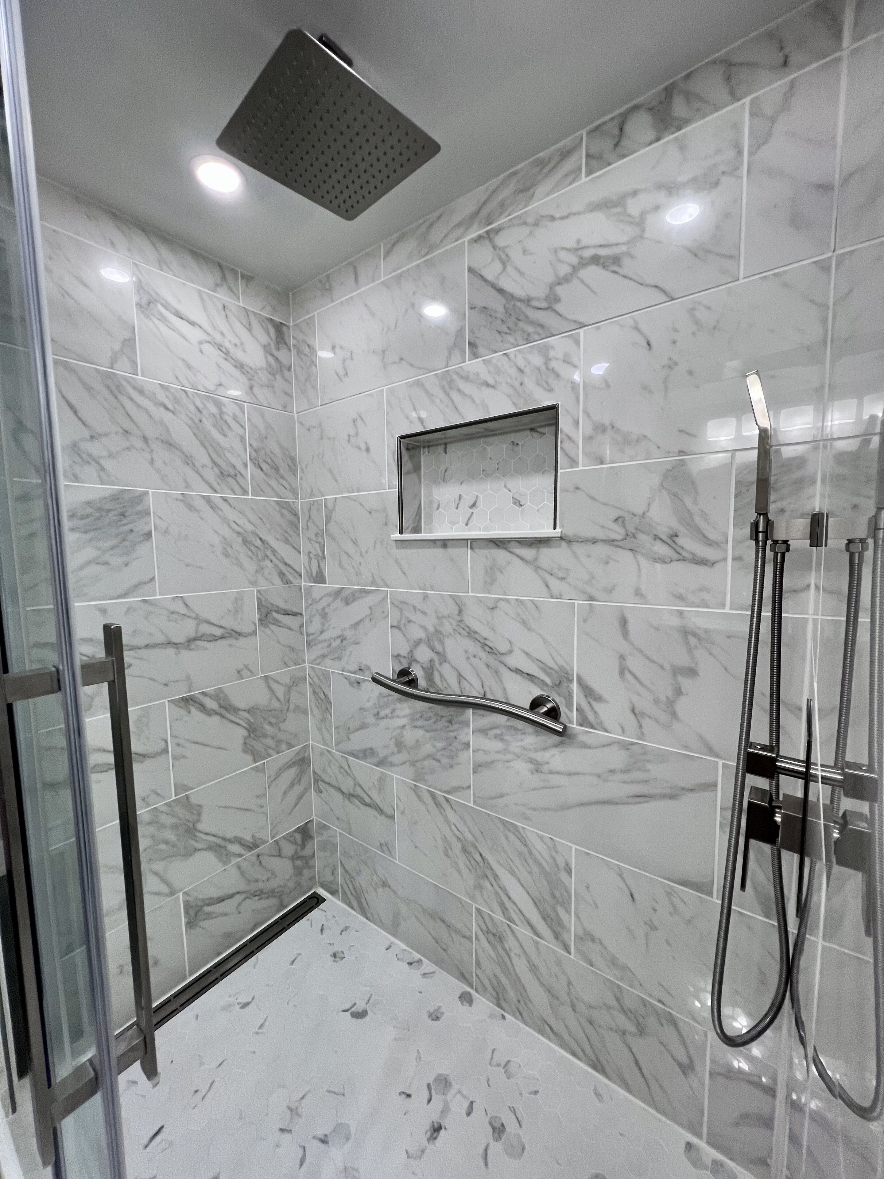 Un espace douche moderne doté d’une porte en verre, d’un pommeau de douche à main et d’une étagère rectangulaire.