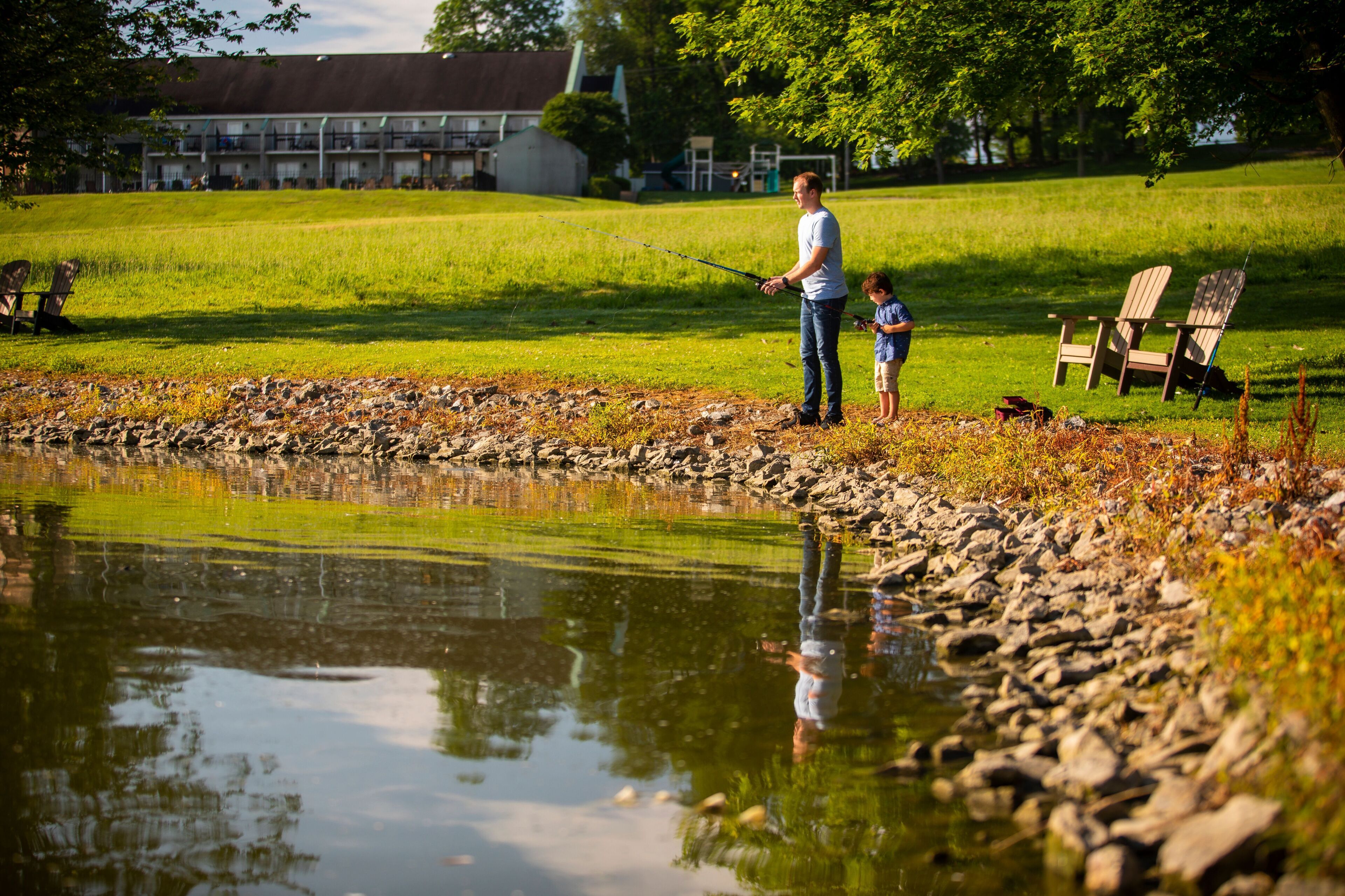 A man and a child fishing by a pond with chairs and a building in the background.