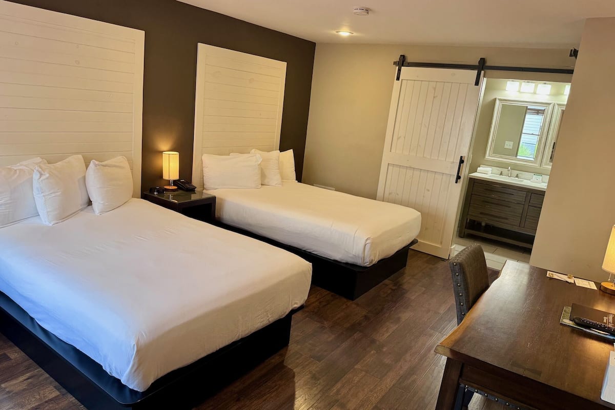 A hotel room with two beds, a desk, a nightstand, a lamp, and a bathroom visible through an open door.