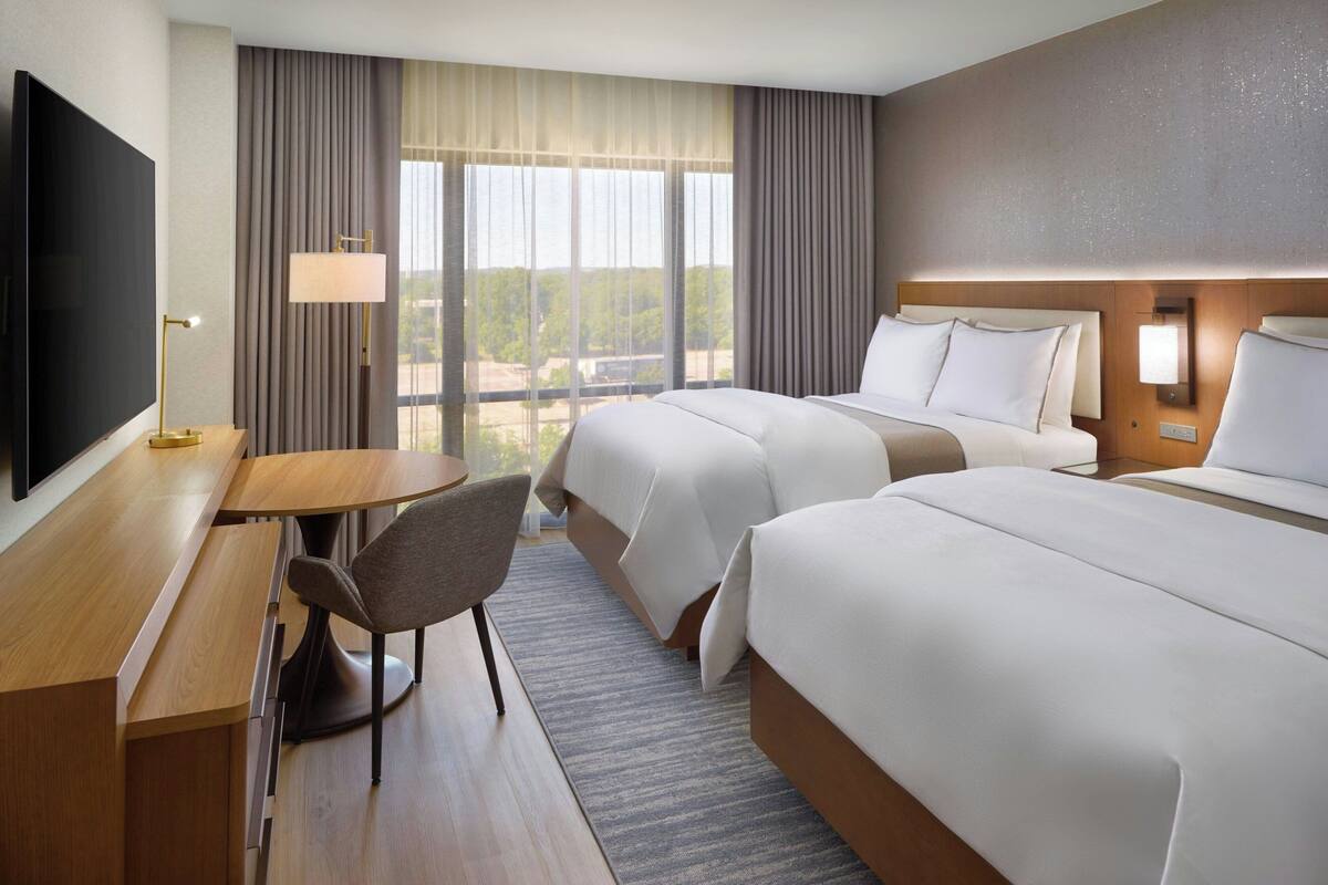 Een hotelkamer met twee bedden, een bureau, een stoel, een televisie en een groot raam met gordijnen.