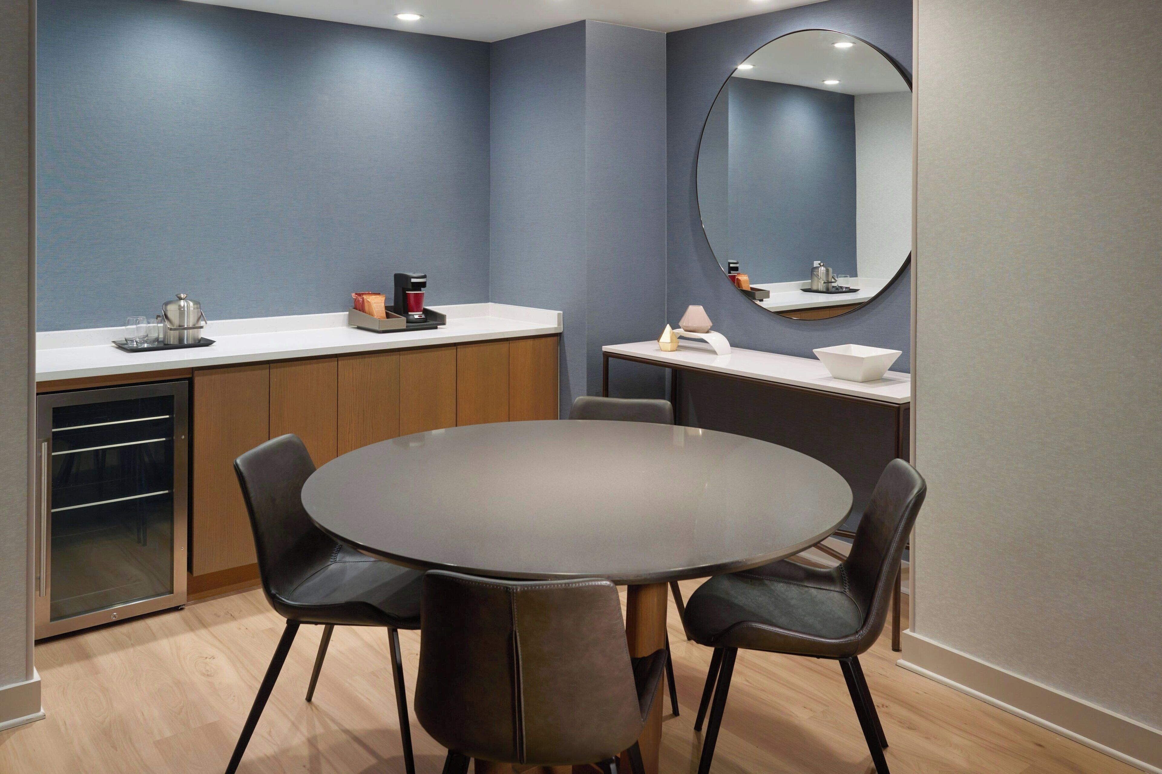 A modern kitchen with a round dining table, black chairs, a coffee machine, and a refrigerator.