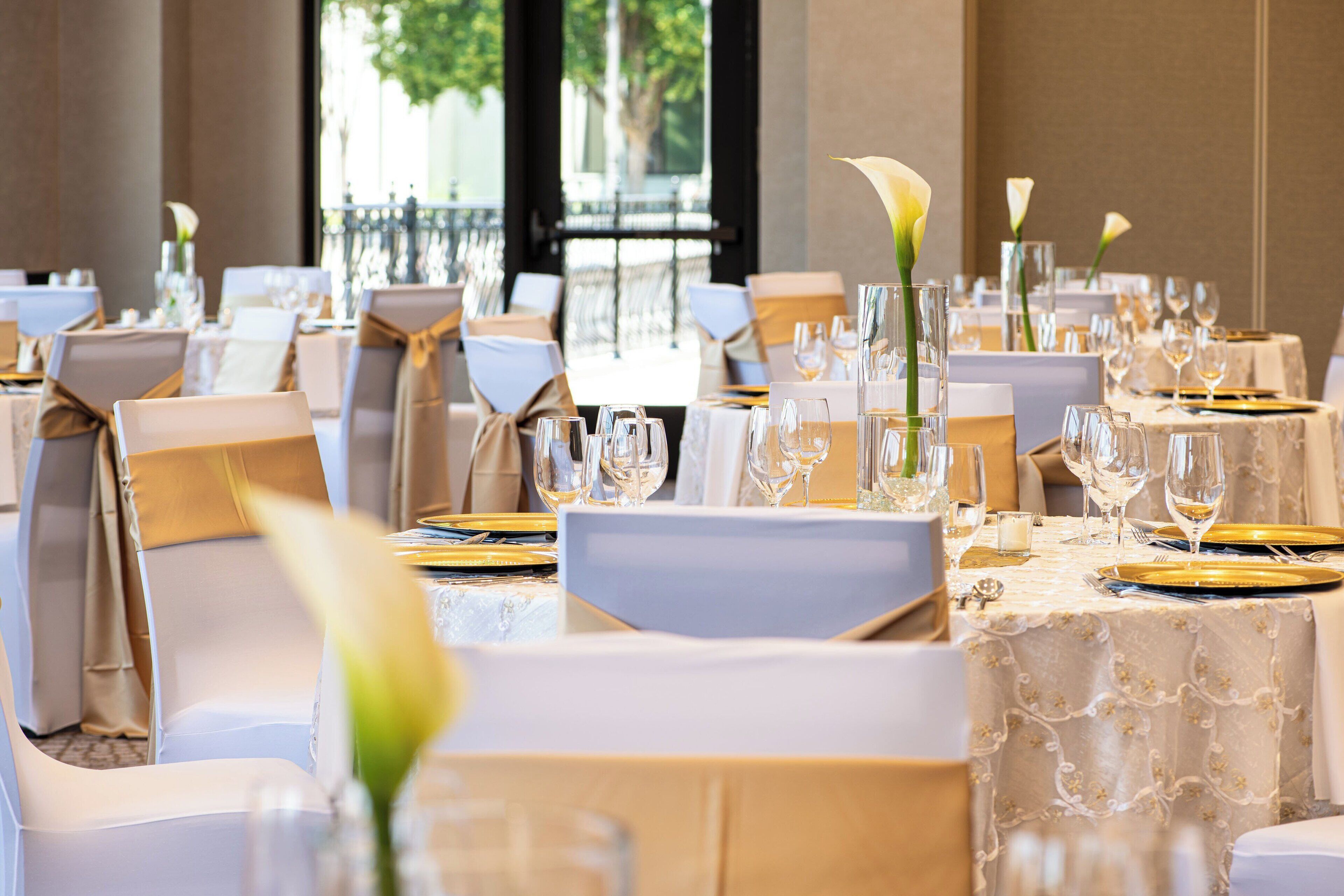 A banquet hall with tables set for a formal event, featuring white tablecloths, gold chargers, and floral centerpieces.