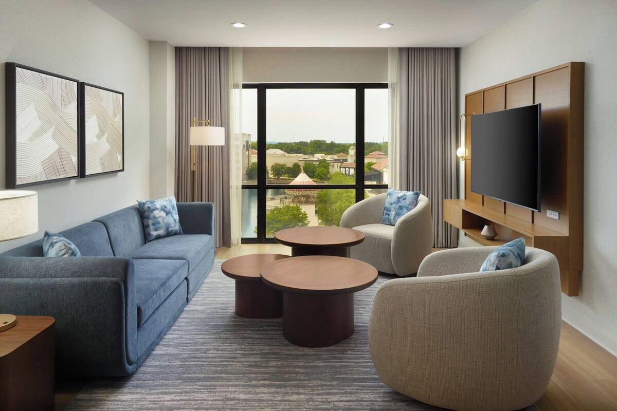 Een moderne hotelkamer met een bank, fauteuil en een rond bijzettafeltje. Een flatscreen-tv is aan de muur bevestigd, en er is een groot raam met uitzicht op de stad.