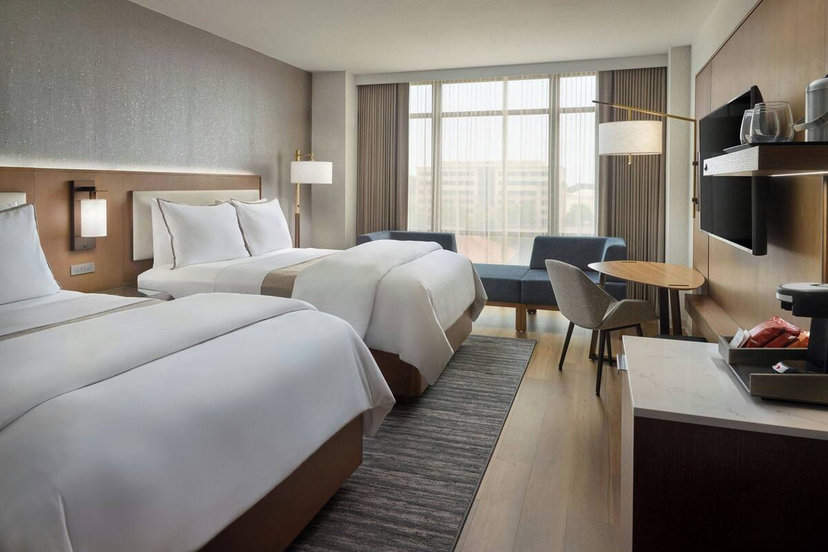 Hotelkamer met twee bedden, een bureau, een stoel, een televisie en een groot raam met gordijnen.