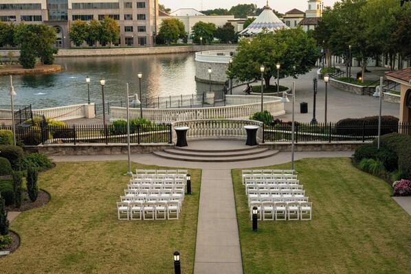 Un lieu de réception en plein air pour les mariages, avec des chaises blanches disposées en rangées face à une scène centrale, le tout entouré d’un mur de pierre et bordé par un plan d’eau.