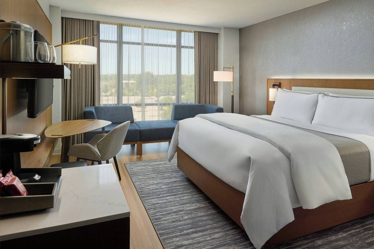 Hotelkamer met een groot bed, een bureau met een computer, een stoel en uitzicht op het omliggende landschap door grote ramen.