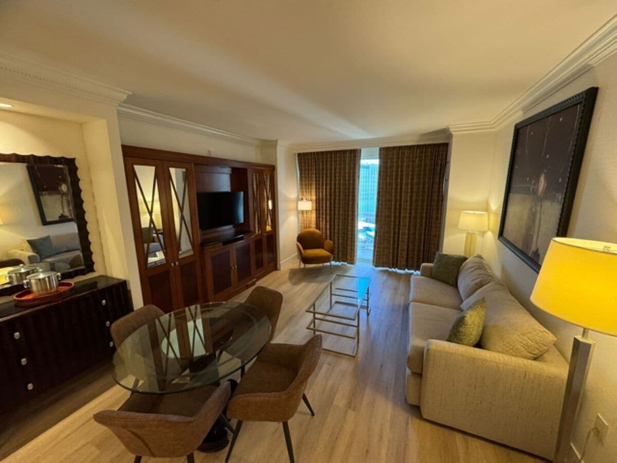 A hotel room with a living area featuring a sofa, armchairs, a glass coffee table, a TV, a wooden cabinet, and a dining table with chairs.