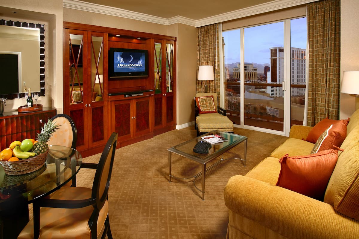 Habitación de hotel con televisión, zona de comedor con frutero, sofá y vista de la ciudad a través de amplias ventanas.