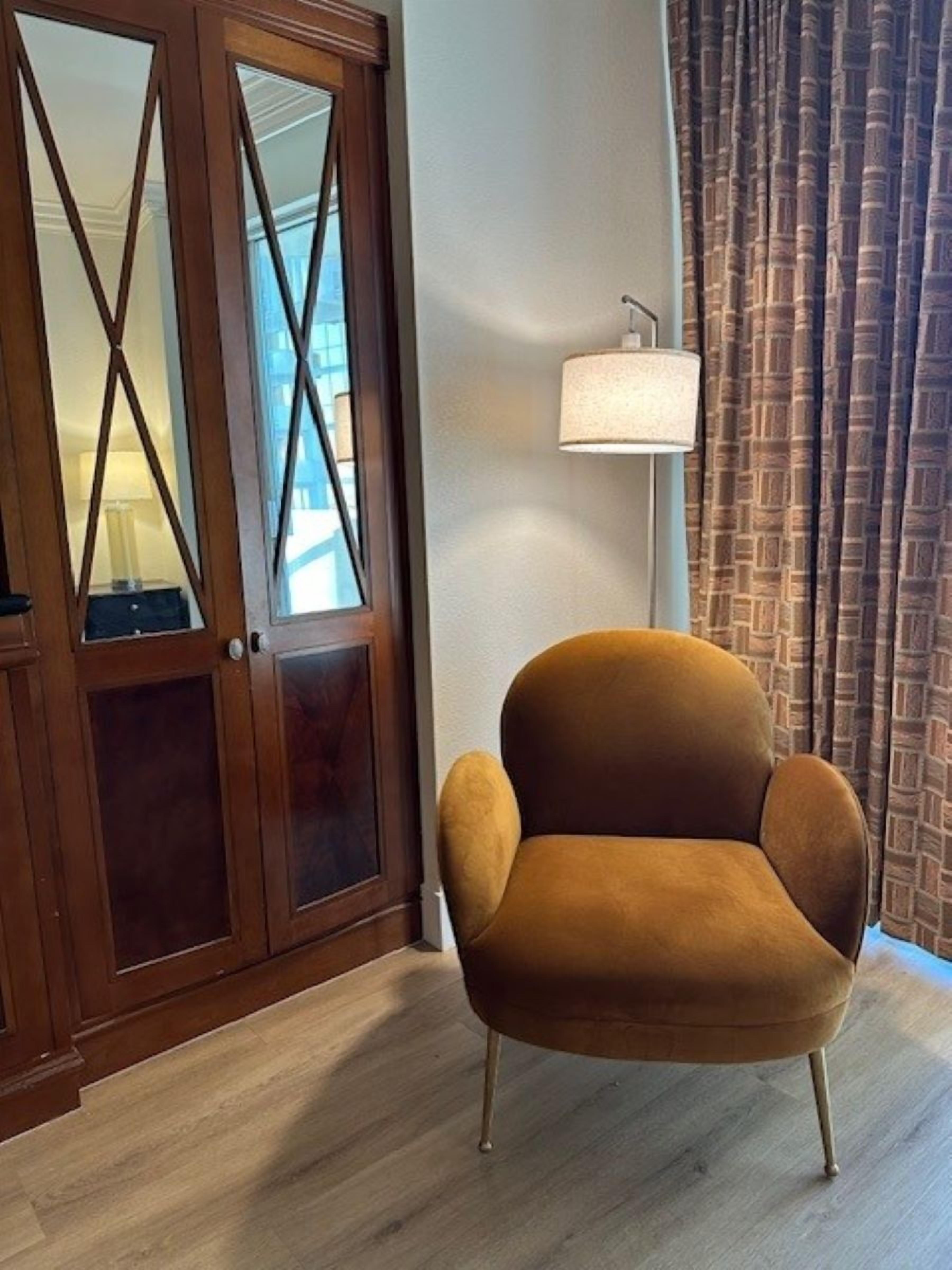 A room with a wooden wardrobe, a brown armchair, a floor lamp, and a patterned curtain.