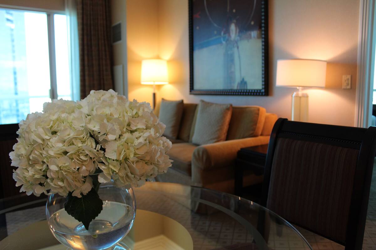 Habitación de hotel con una mesa de vidrio, un florero con flores blancas, un sofá y una lámpara.