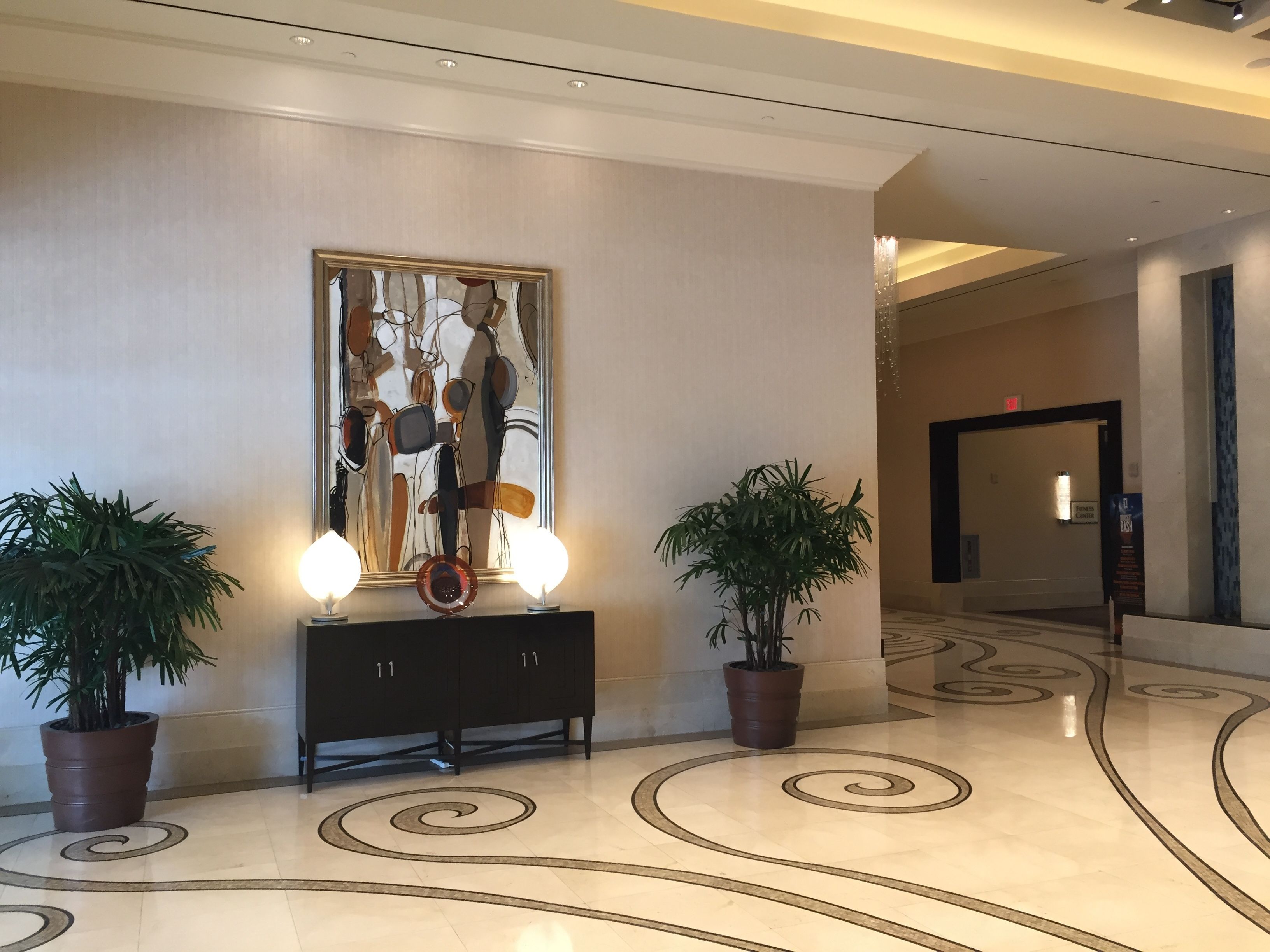 A lobby with a modern design, featuring a large abstract painting, a console table with two lamps, and potted plants.