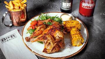 Un plato con pollo a la parrilla, mazorca de maíz y papas fritas, acompañado de botellas de cerveza y una servilleta con el logo "Bier Fabriek".