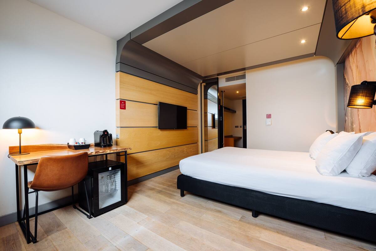A modern hotel room with a bed, a desk with a chair, a coffee machine, and a mini-fridge.