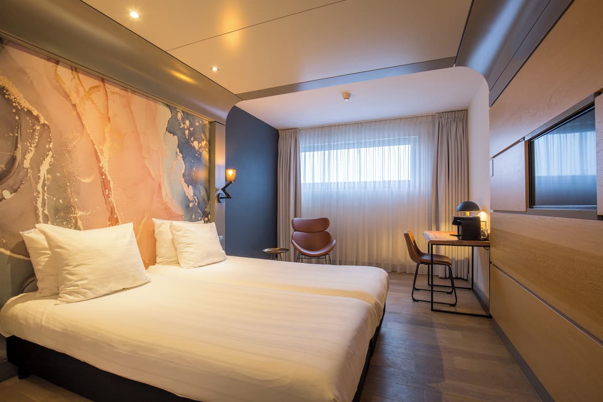 A hotel room with a large bed, a desk with a chair, a small table, and a window with curtains.