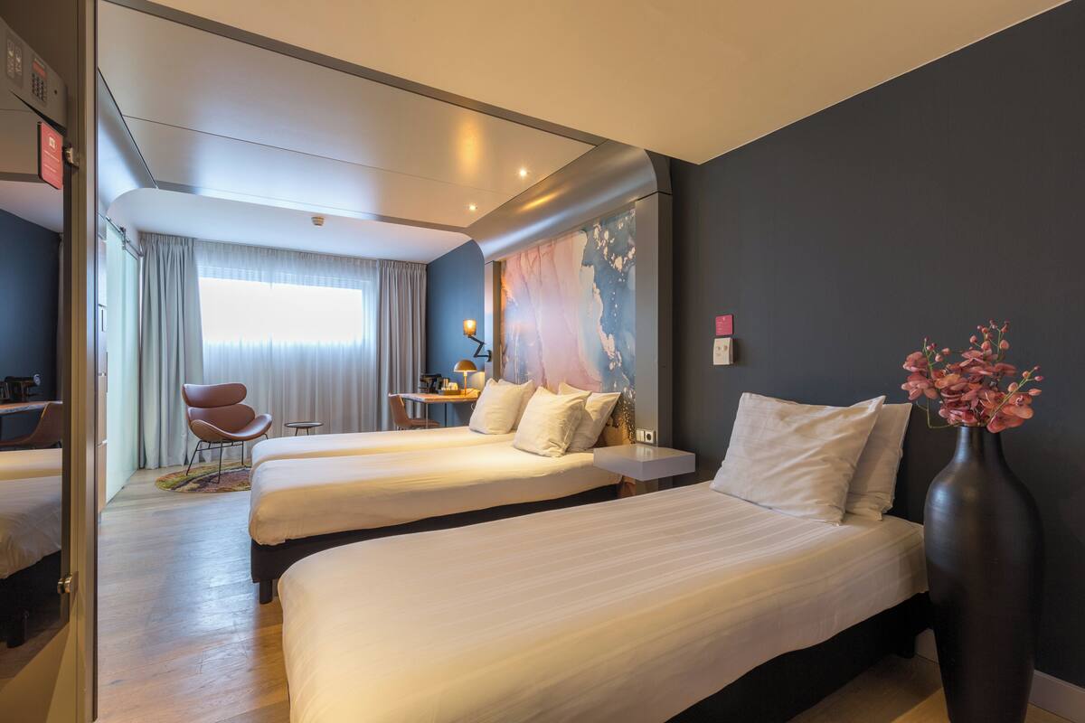 A hotel room with two beds, a desk, a chair, and a large window with curtains.