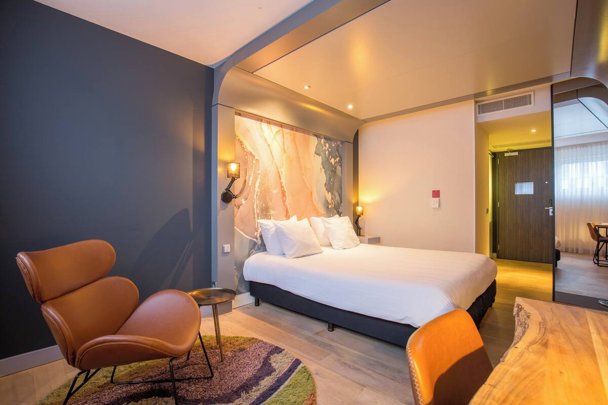 A modern hotel room with a large bed, two armchairs, a small table, and a marble accent wall.