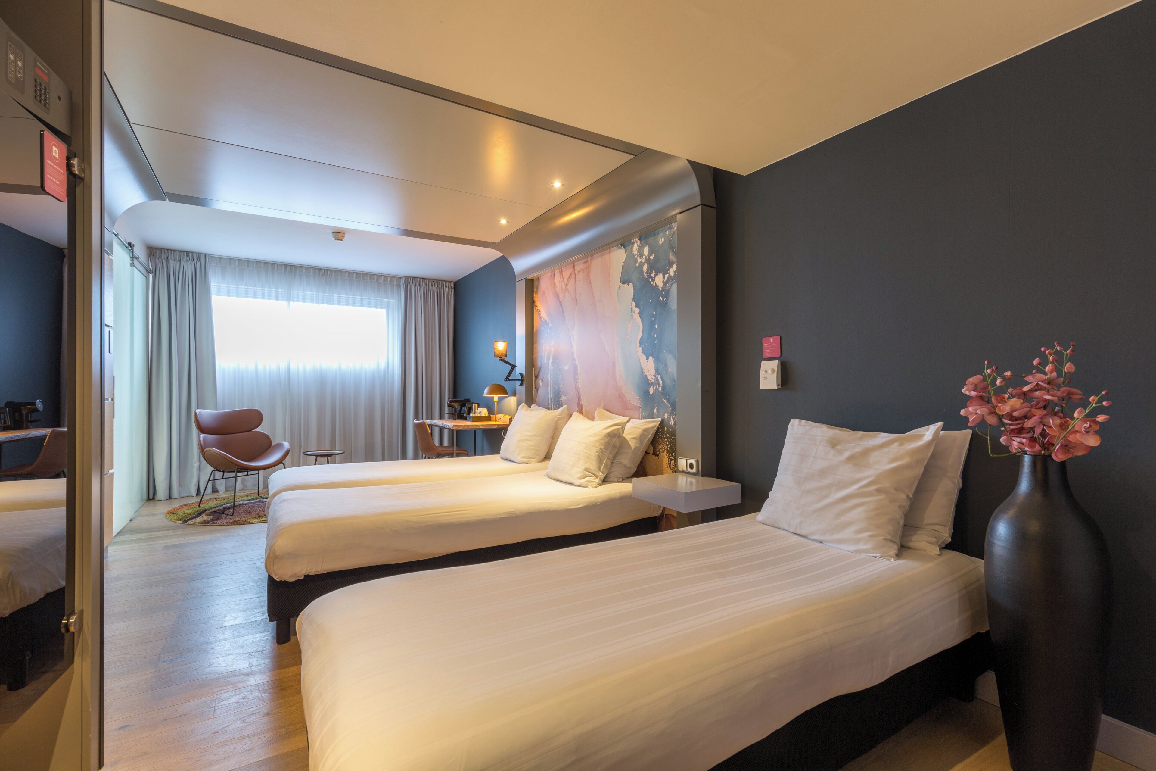 Une chambre d’hôtel avec deux lits, un bureau, une chaise et une grande fenêtre avec des rideaux.