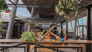 Uma área de refeições ao ar livre com mesas e bancos de madeira, uma placa para BIER FABRIER e plantas penduradas.