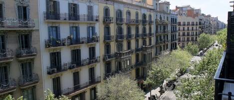 A row of multi-story buildings with balconies, a street with parked scooters, and trees.