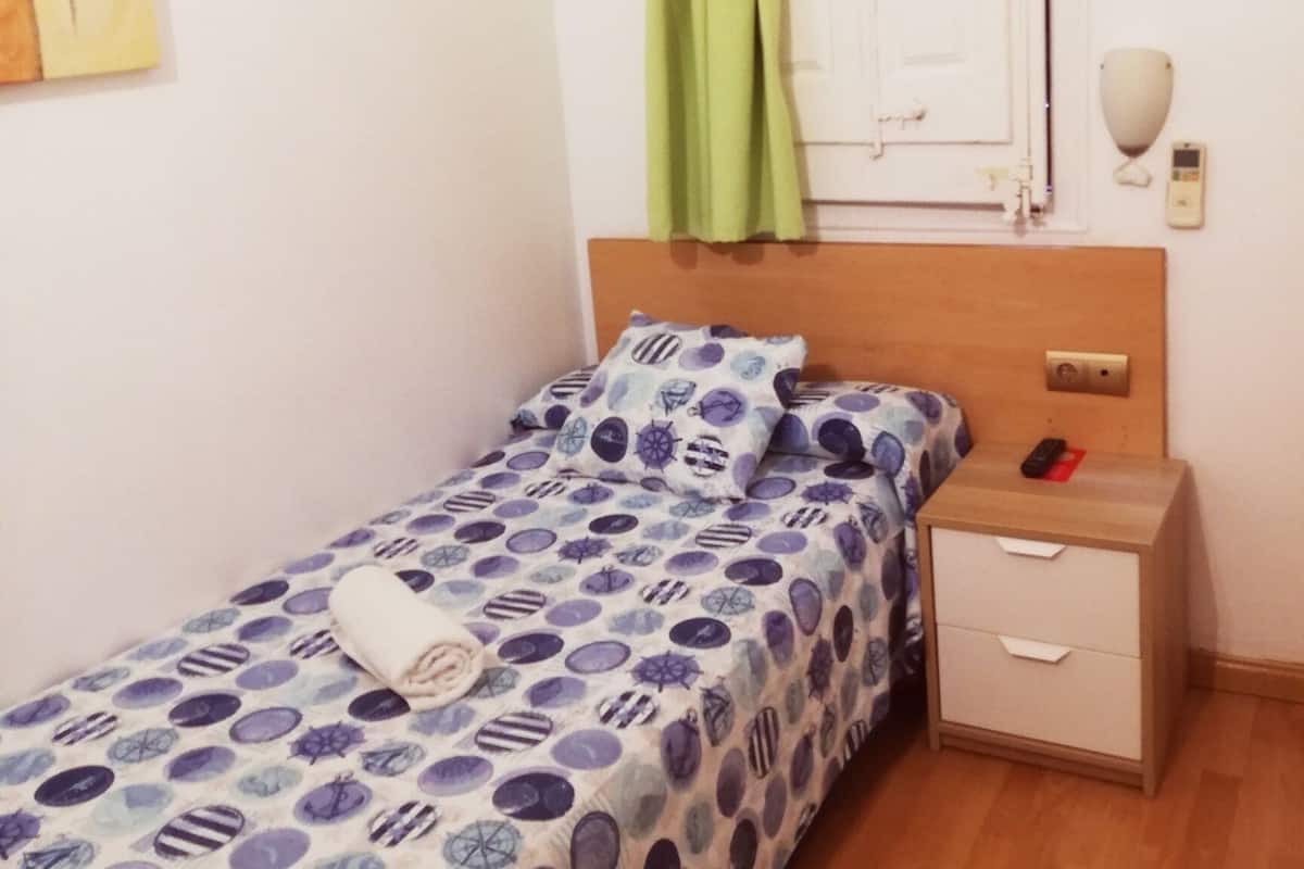 Una cama individual con una colcha estampada, una mesita de noche con un teléfono y una cortina verde.