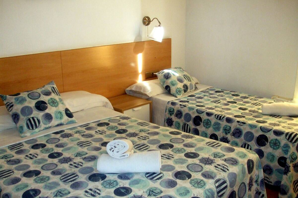 Habitación de hotel con dos camas, cada una con colcha estampada, cabecero de madera, mesita de noche, lámpara y luz en la pared.