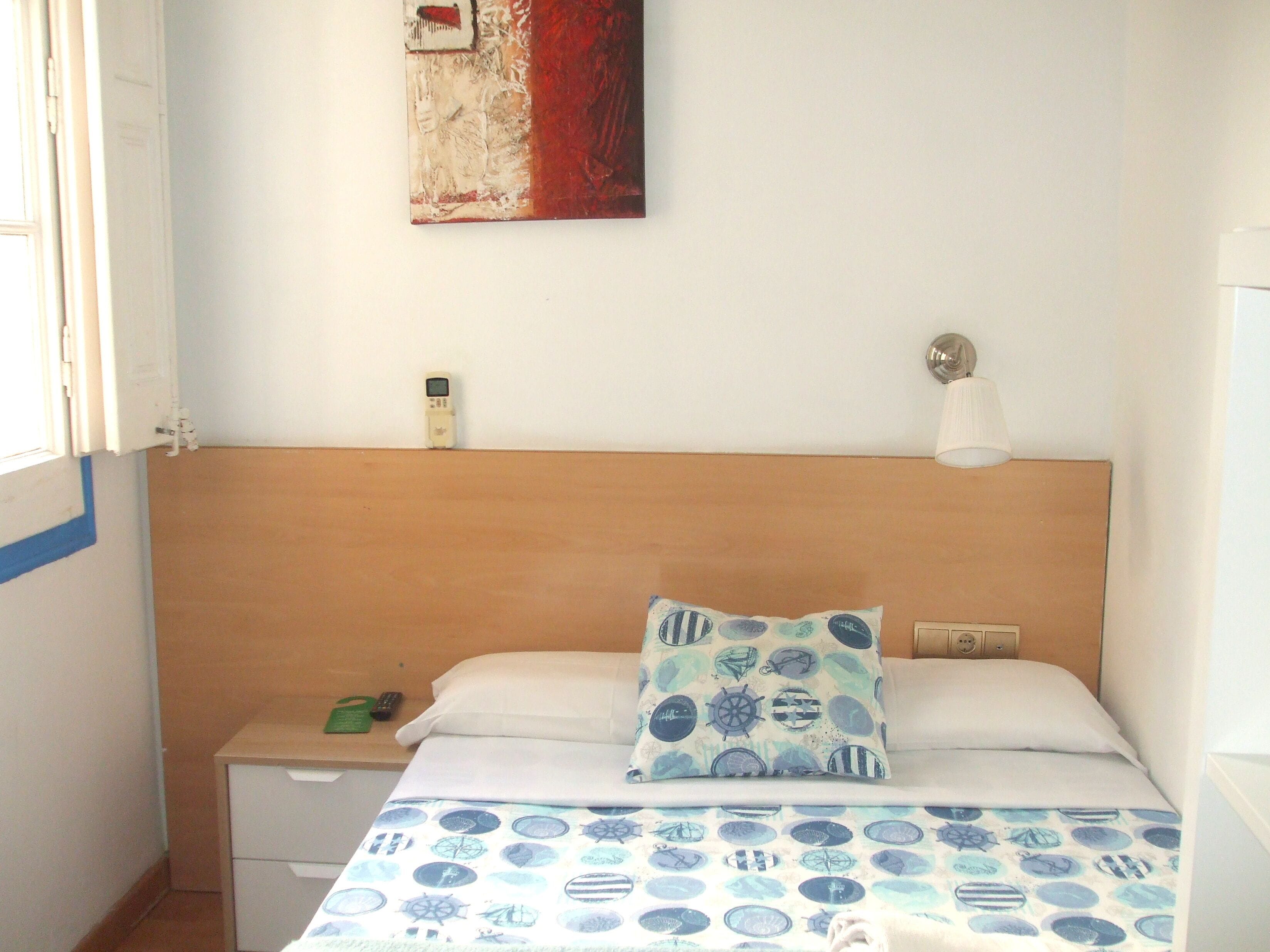 Une chambre à coucher avec une tête de lit en bois, un lit recouvert d’une couvre-lit à motifs, une table de chevet sur laquelle se trouve un objet vert, et une lampe fixée au mur.