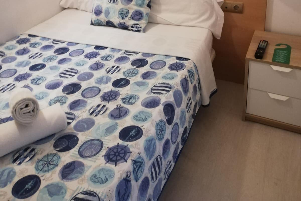 Una cama bien hecha con una colcha estampada, cabecera de madera, lámpara de noche, mesita de noche con control remoto y teléfono fijo en la pared.
