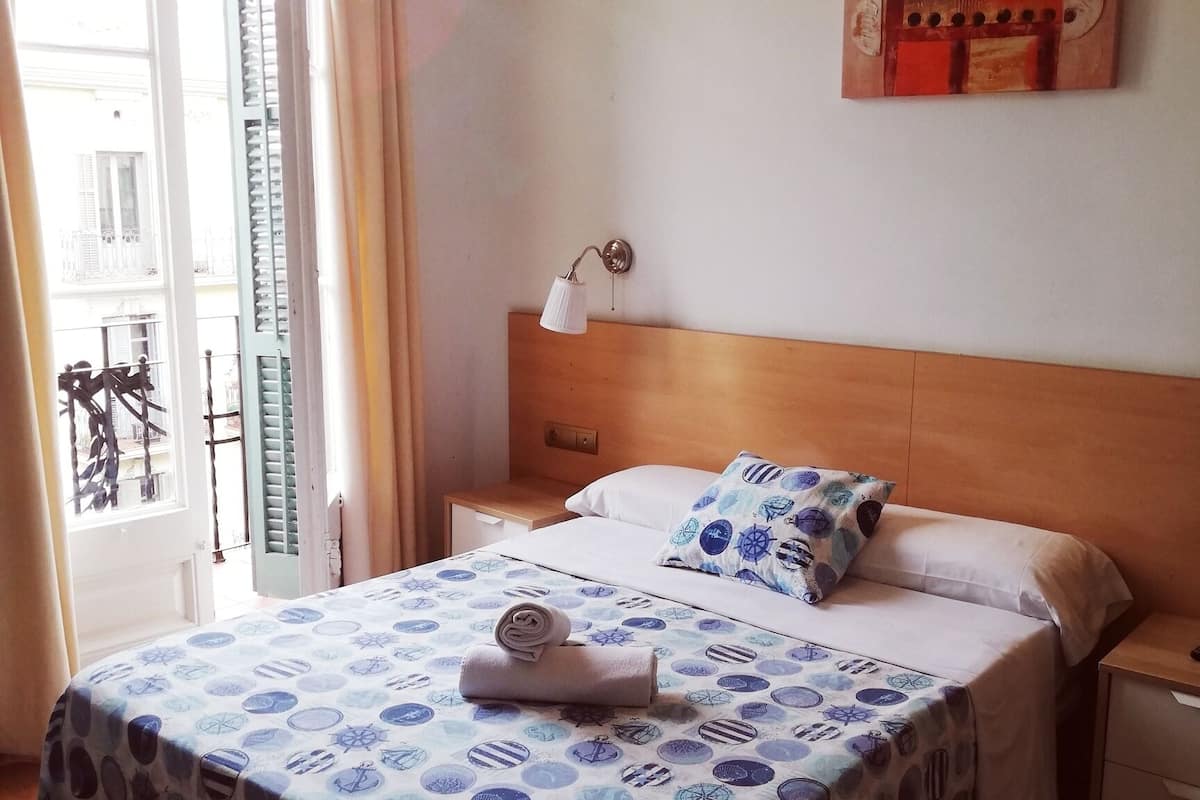 Una habitación de hotel con una cama, una mesita de noche, una lámpara de pared y un ventanal con cortinas.