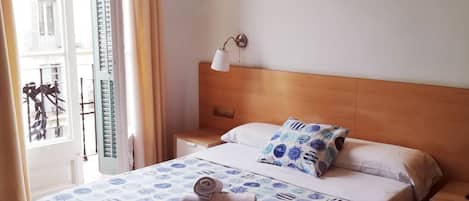 Una habitación de hotel con una cama, una mesita de noche, una lámpara de pared y un ventanal con cortinas.