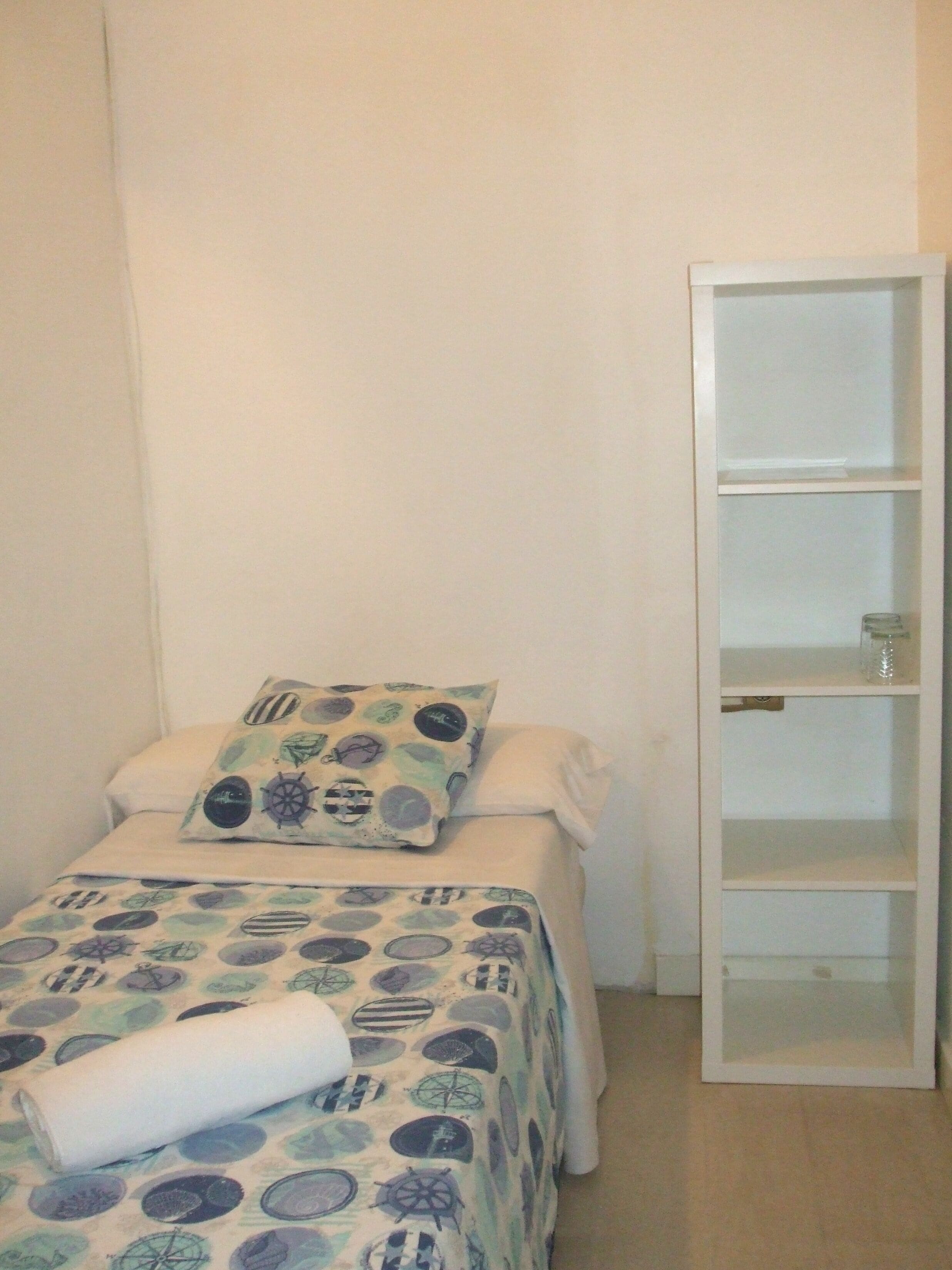 Een klein, netjes opgemaakt bed met een bedsprei met dessin, een kussen, een witte nachtkastje met een glas, en een witte garderobe.