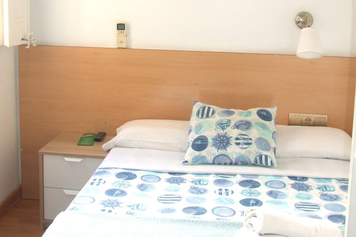 Una habitación de hotel con cama, mesita de noche, un cuadro en la pared, lámpara y ventana.