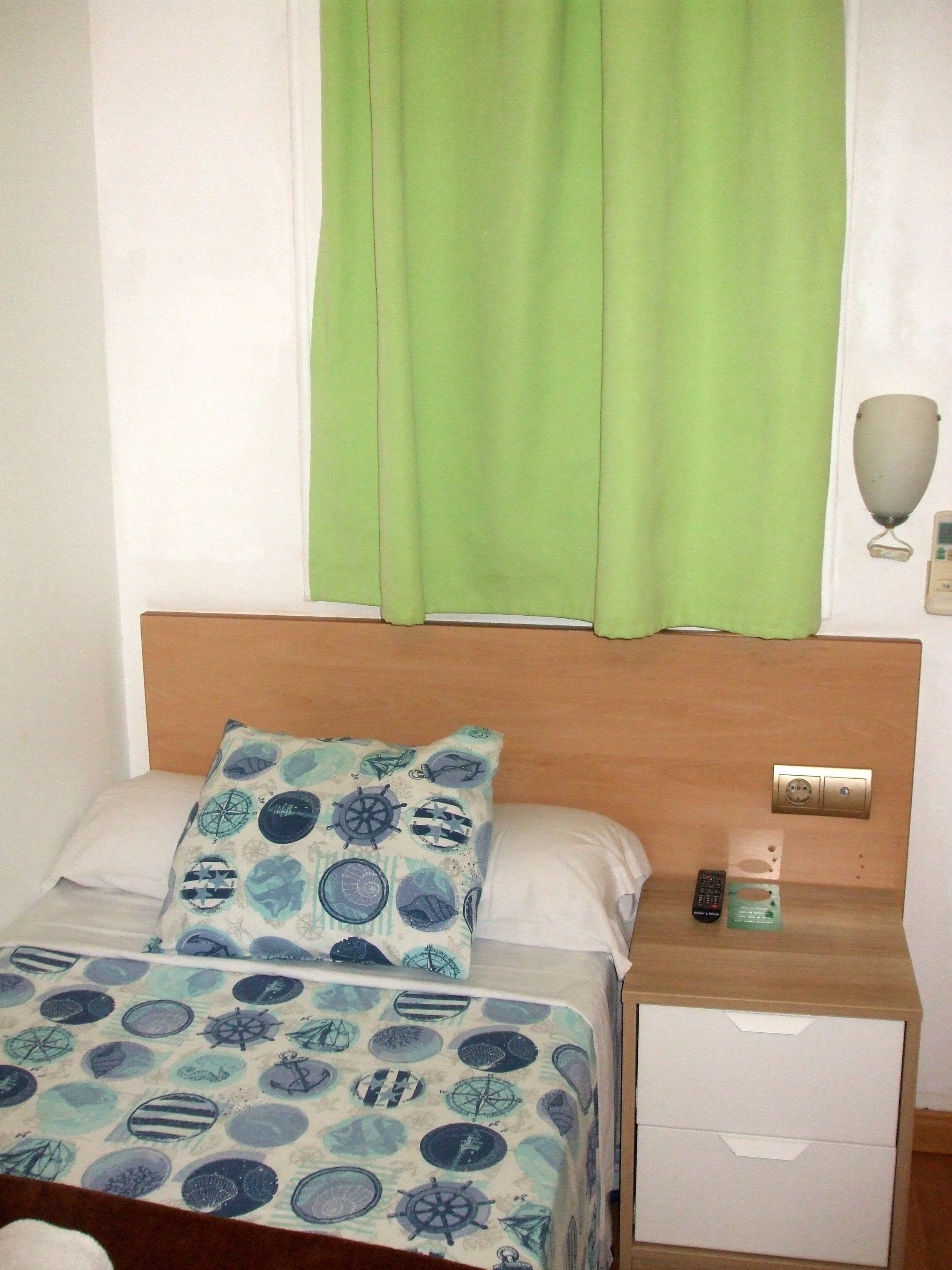A hotel room with a bed, a bedside table with a remote control, and a nightstand with a lamp.