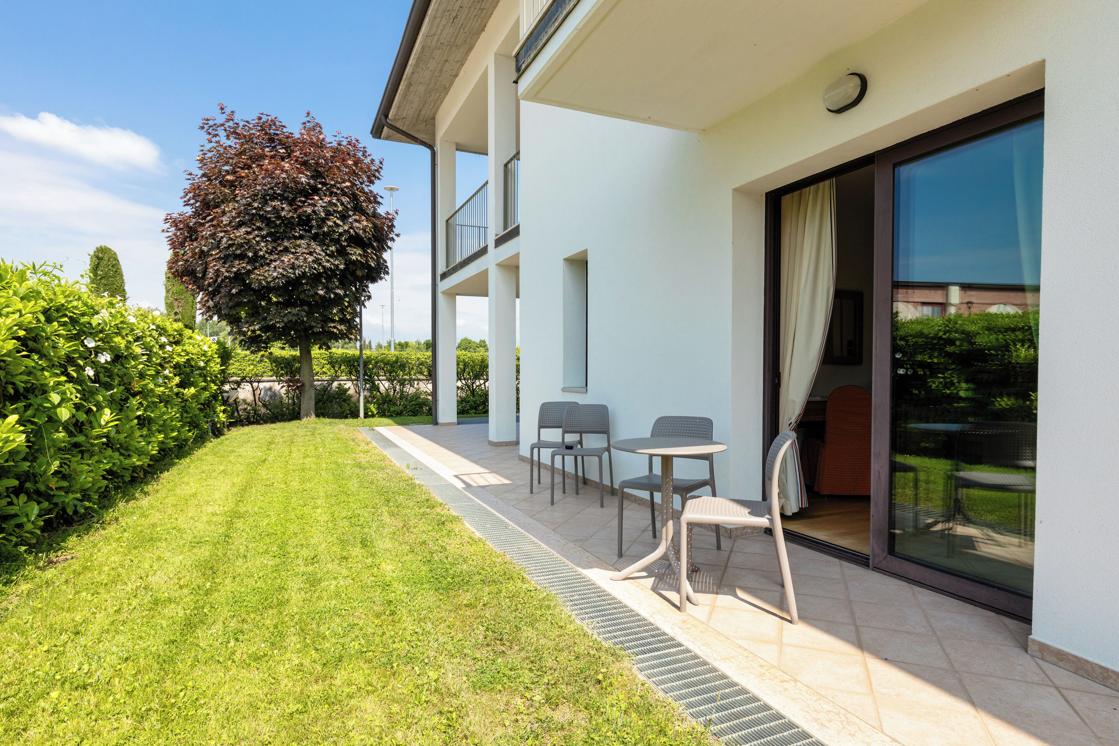 Un patio exterior moderno con césped cuidado, un árbol y una zona de estar con sillas y una mesa.