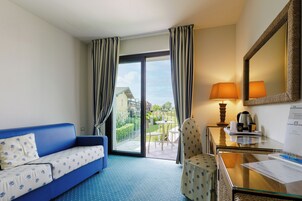 Habitación de hotel con sofá azul, una mesita con lámpara, una silla y un balcón con vistas a edificios y zonas verdes.
