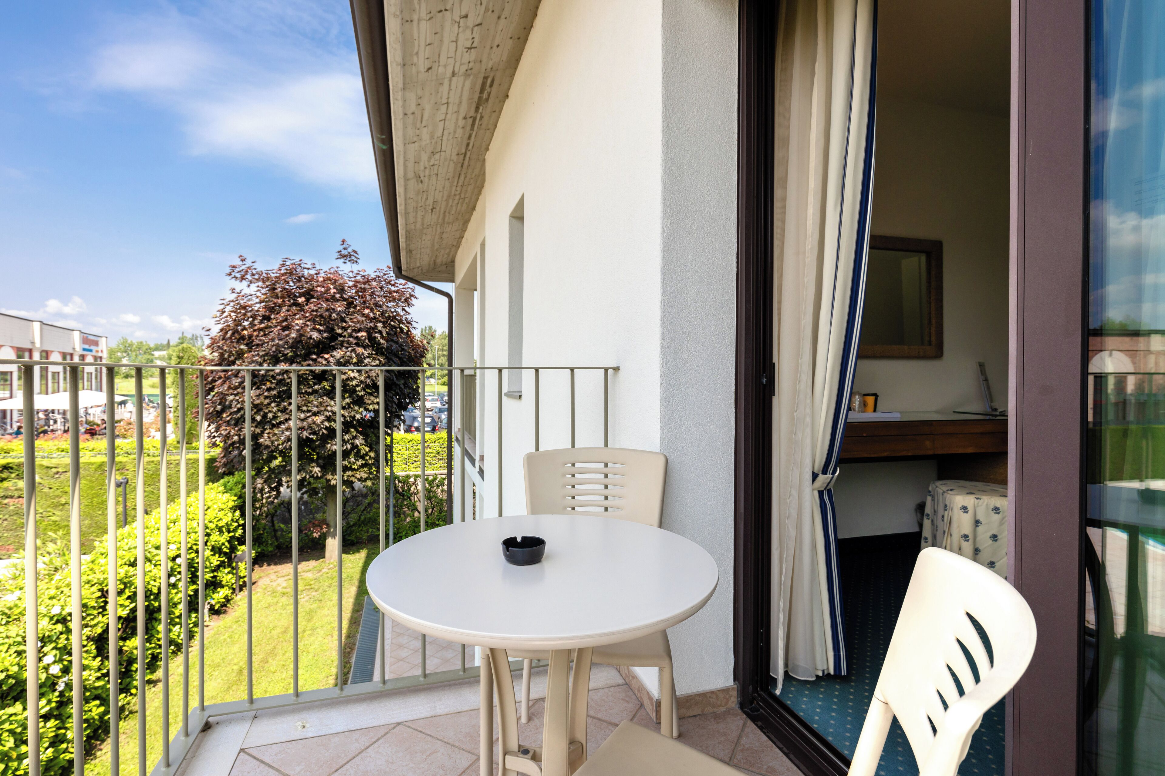 En balkon med et rundt bord og to stole, udsigt over grønt og en bygning i baggrunden.