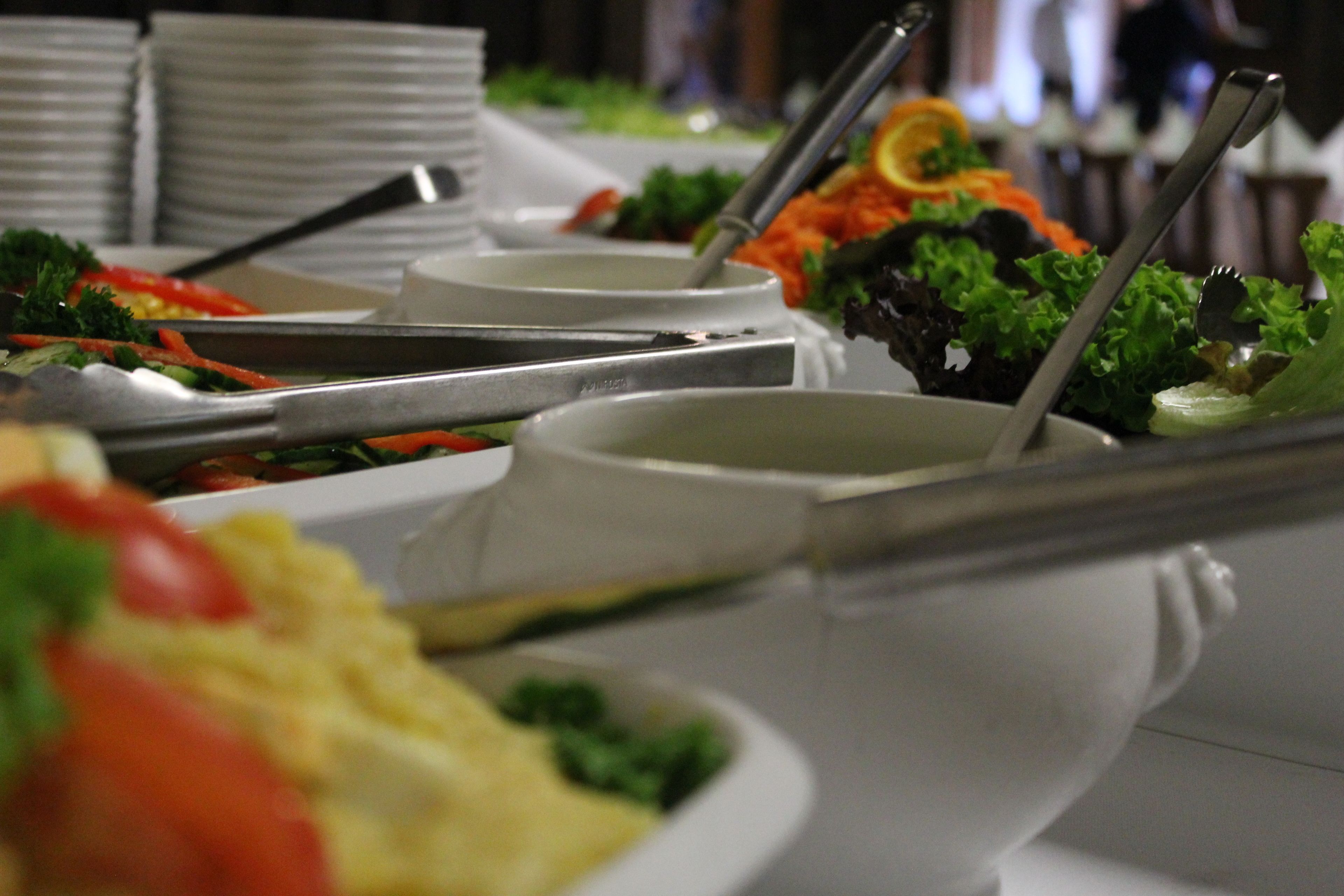 Buffet com diversos pratos, incluindo saladas e legumes, dispostos em pratos brancos e talheres.