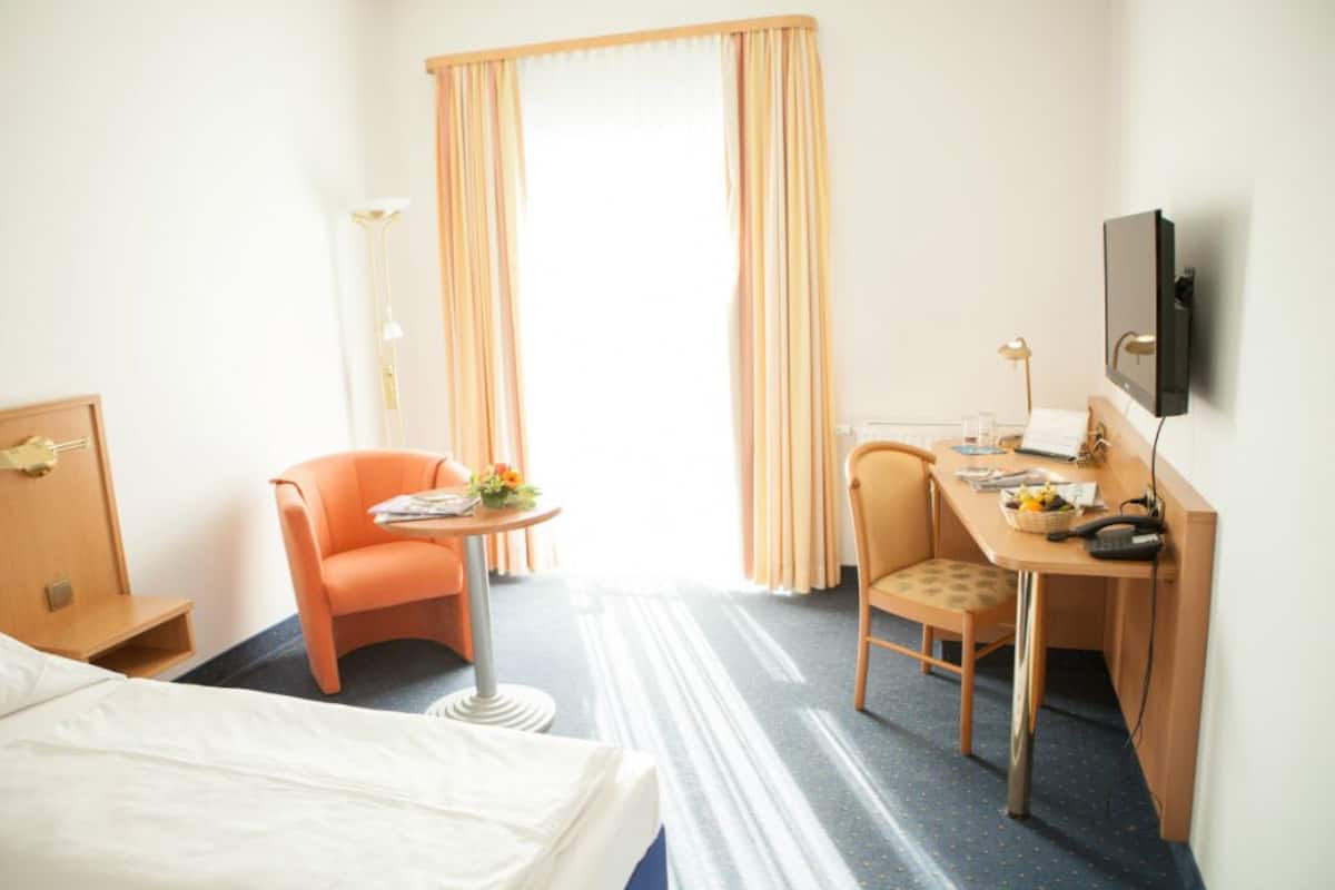 Une chambre d’hôtel comprenant un lit, un bureau, deux chaises, une télévision et une fenêtre avec des rideaux.