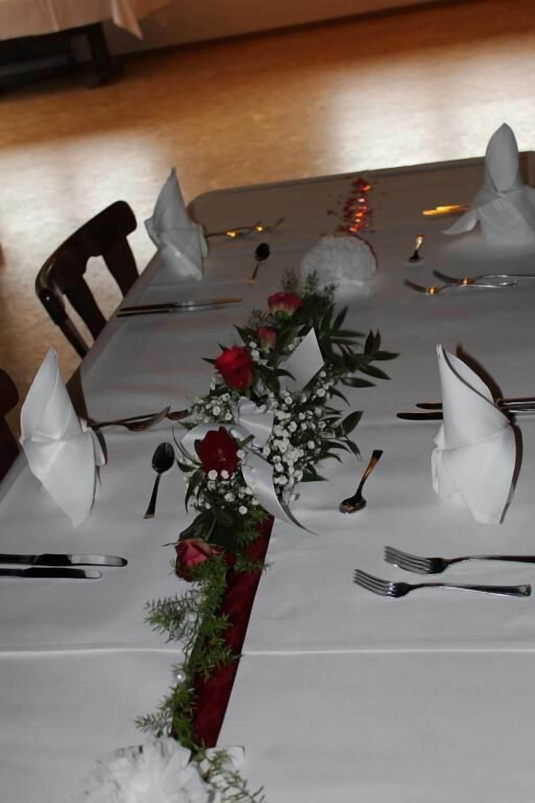 Ein Tisch ist für eine festliche Veranstaltung gedeckt, mit weißen Tischdecken, gefalteten Servietten und einer Blumendekoration aus roten Rosen und Grünpflanzen.