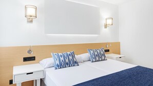 Una habitación de hotel moderna con cabecera de madera, dos mesitas de noche, una cama grande con ropa de cama blanca y almohadas estampadas en azul, y lámparas fijadas a la pared.