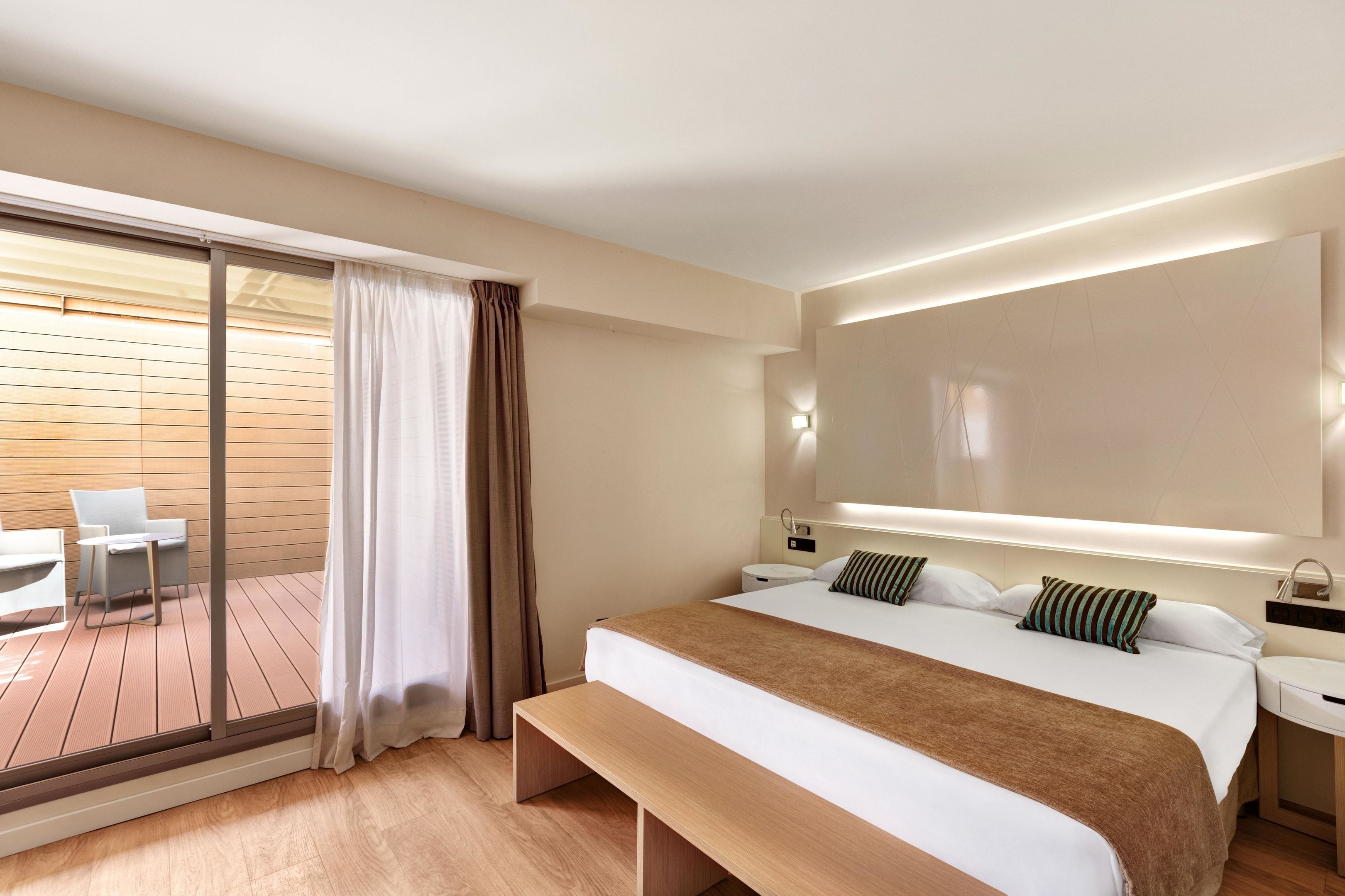 Una habitación de hotel moderna con una cama grande, un banco, un escritorio y un balcón con mesa y sillas.