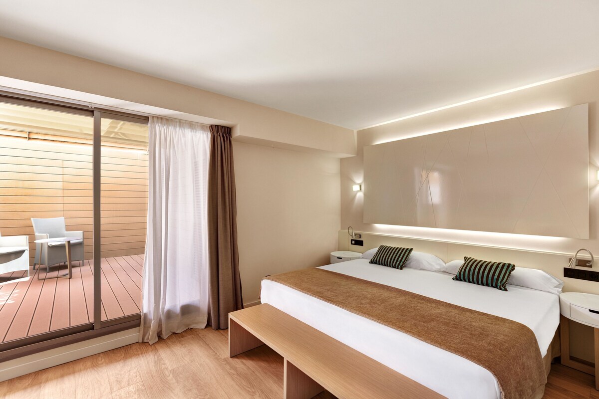 Una habitación de hotel moderna con una cama grande, un banco, un escritorio y un balcón con mesa y sillas.