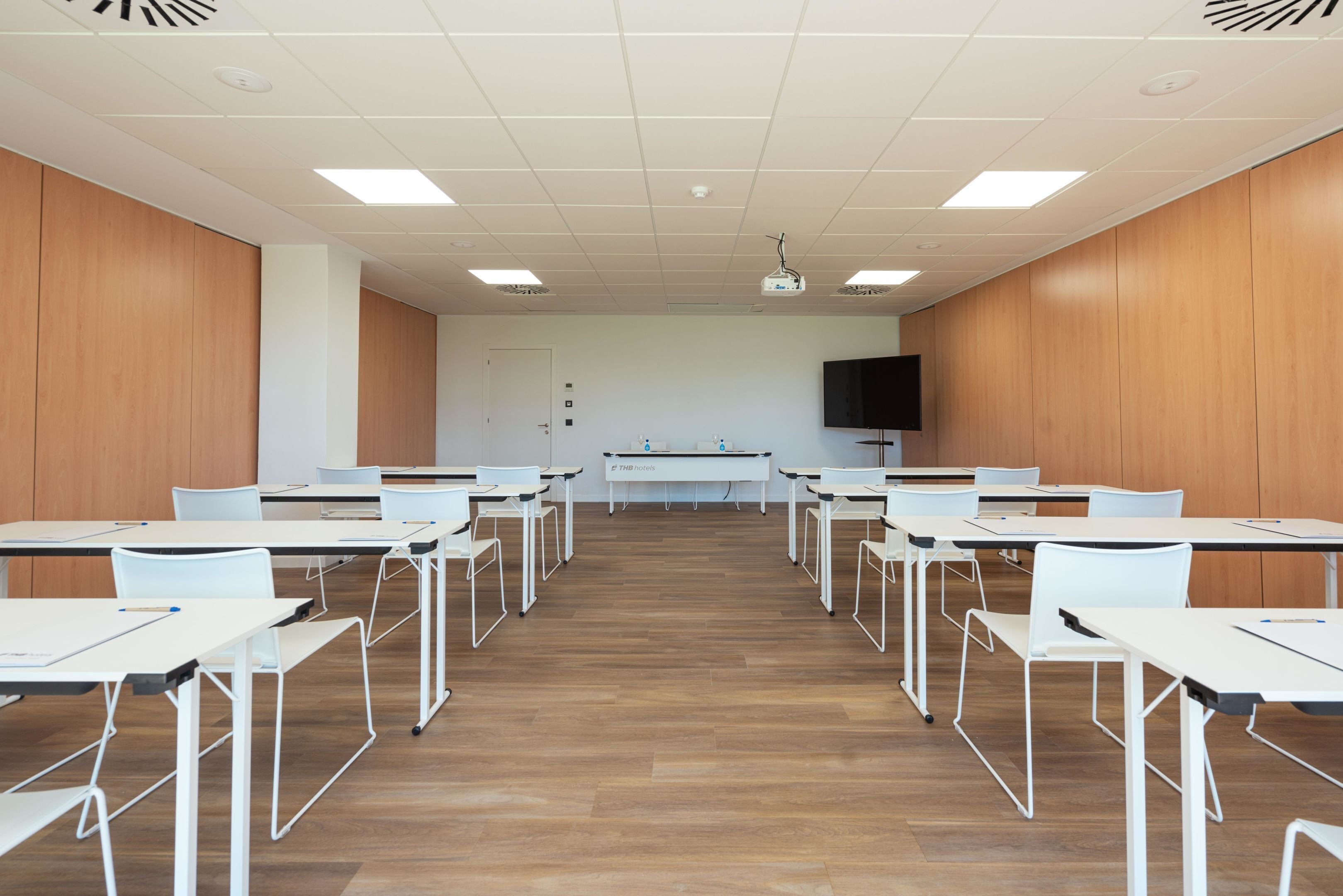 A conference room with white tables and chairs, a projector, and a TV on the wall.
