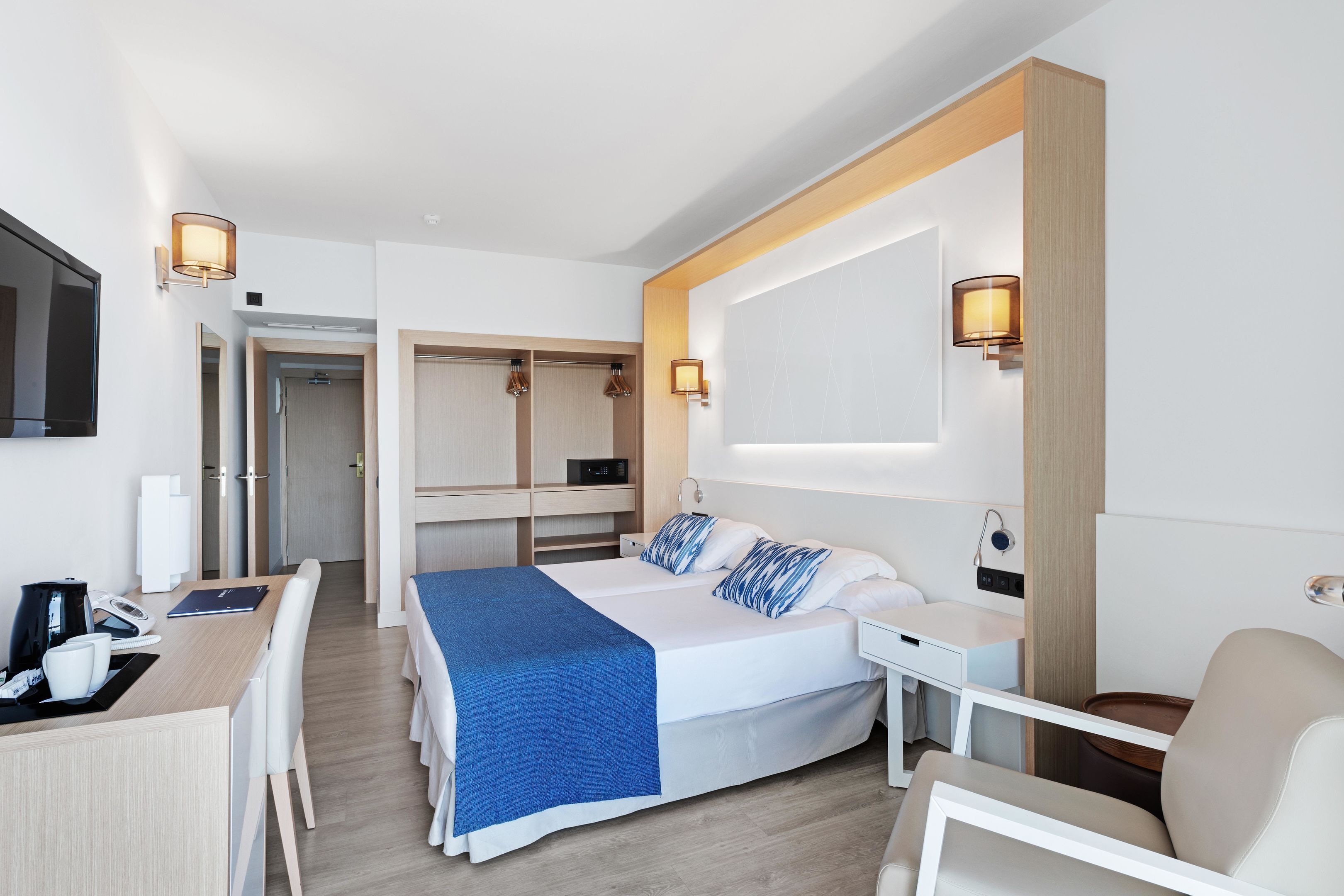 A modern hotel room with a bed, desk, and chair, a small table with a lamp, and a TV mounted on the wall.