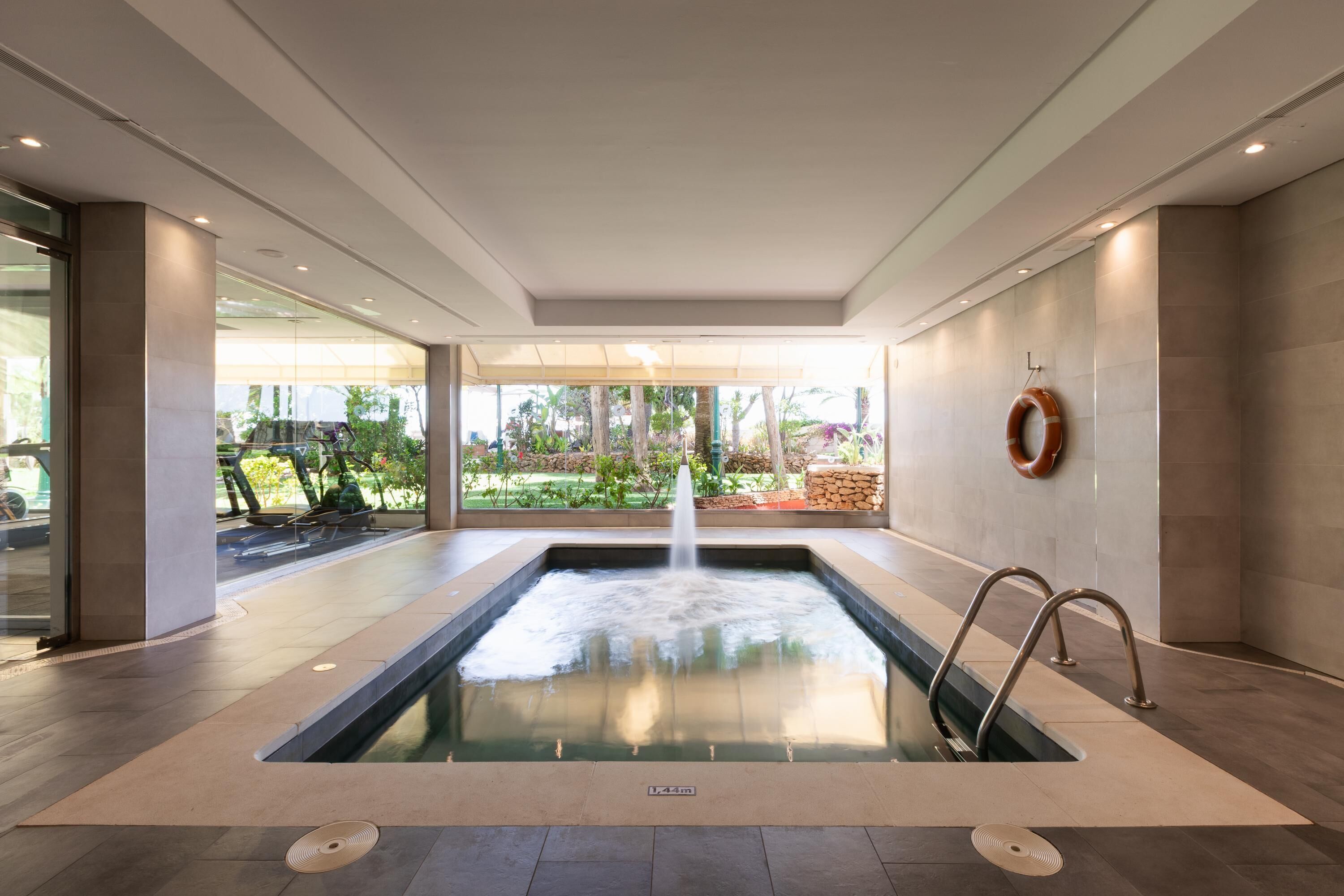Ein moderner Innenpool mit einem zentralen Wasserspiel, umgeben von einem geräumigen Bereich mit Blick auf einen Garten und Außenmöbel.