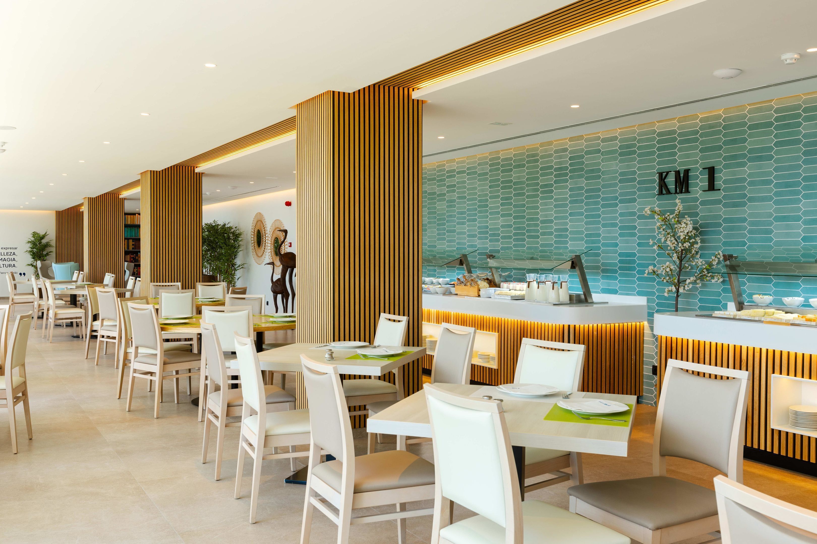 Un restaurant moderne avec des tables et des chaises blanches, un comptoir dont le mur arrière est recouvert de carreaux bleus, et une enseigne indiquant « KM 1 ».