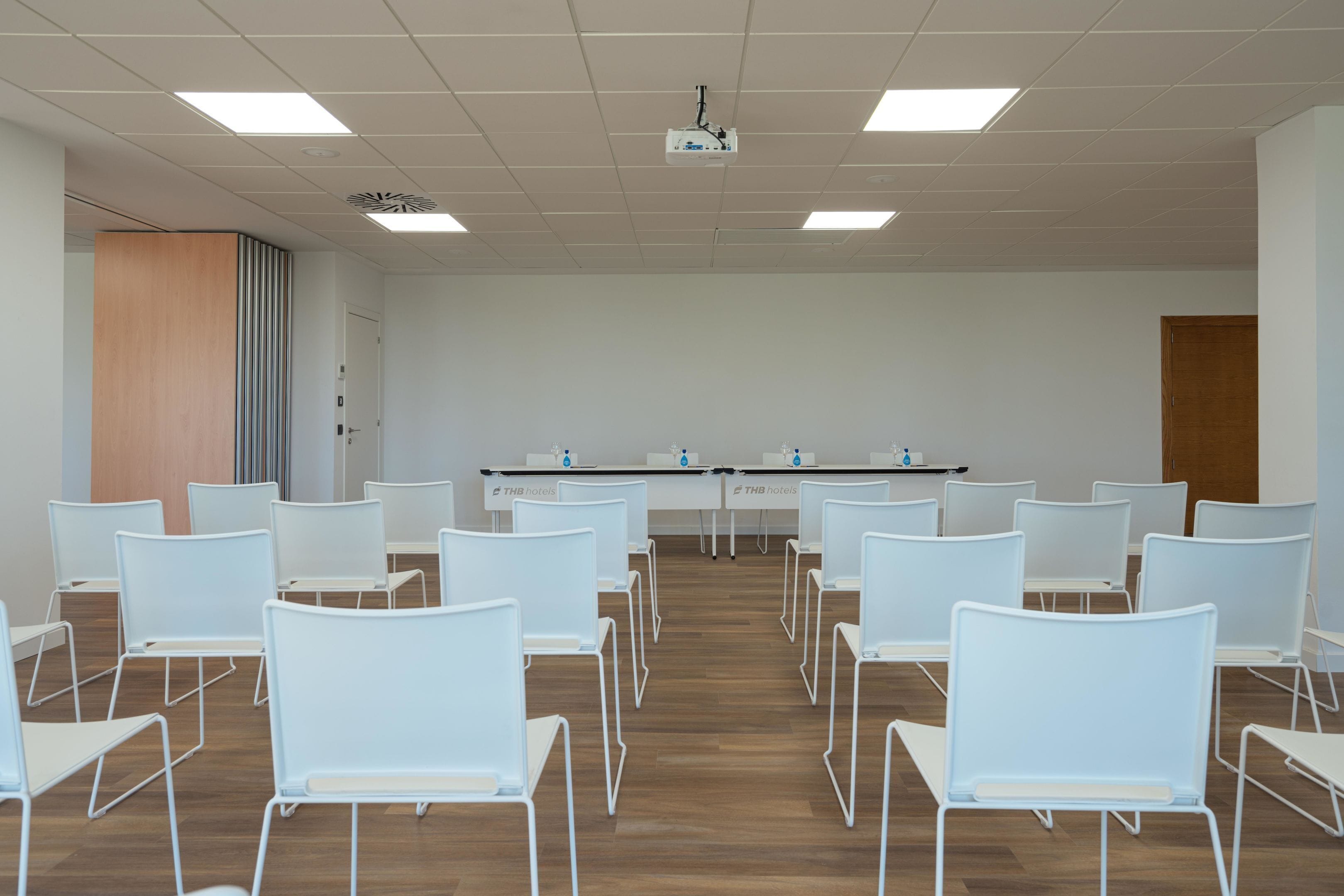 A conference room with white chairs arranged in rows, a projector on the ceiling, and a table with a THB hotels logo.