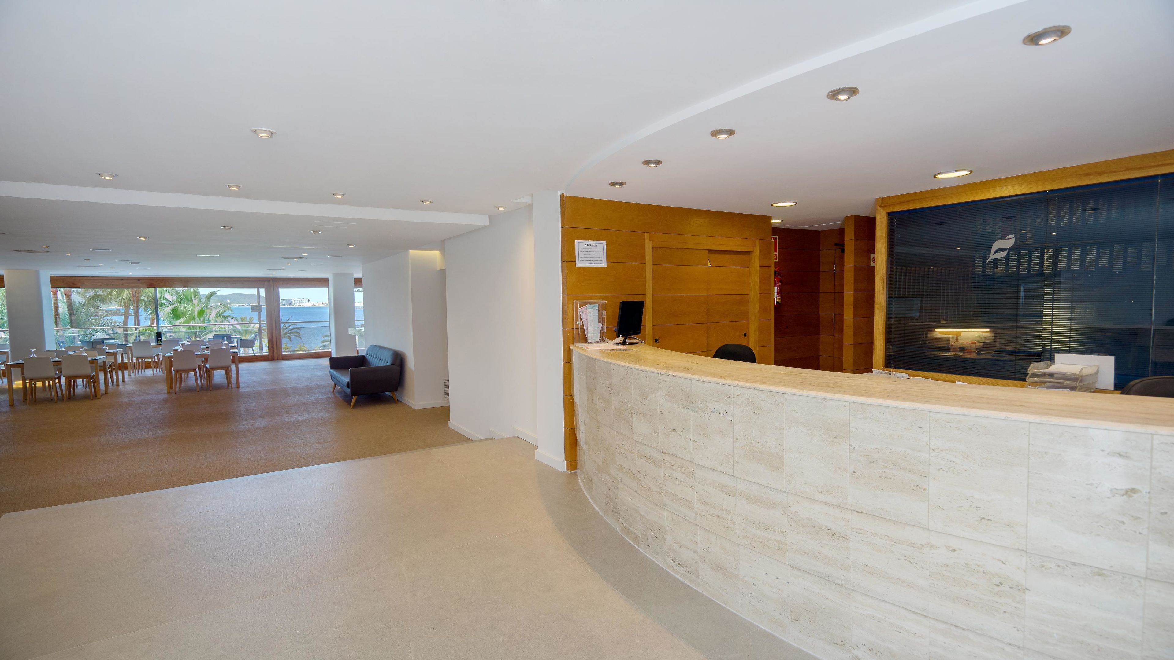 Een hotel receptie met een gebogen receptiebalie, een zithoek met tafels en stoelen, en uitzicht op palmbomen door grote ramen.