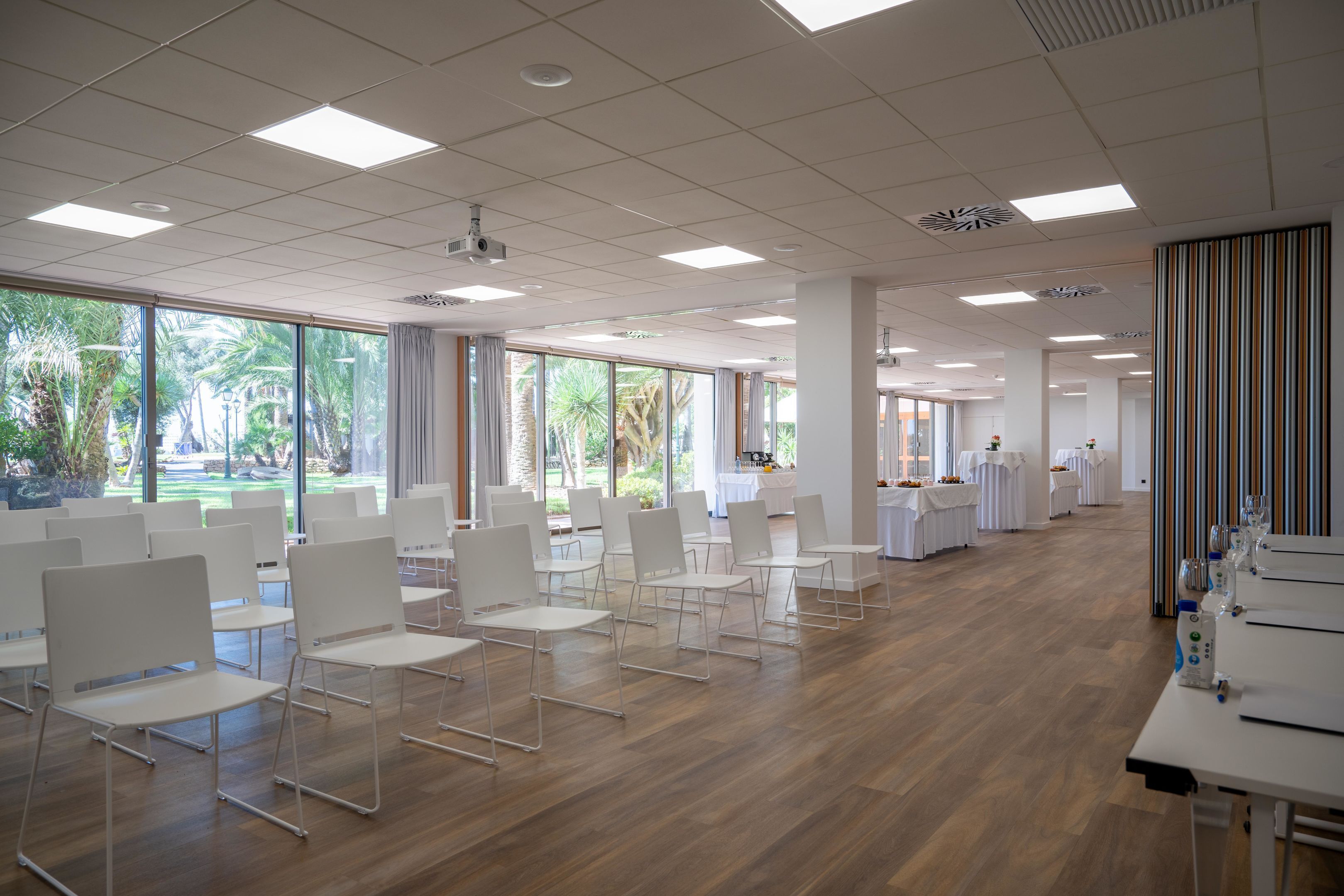Una amplia sala de conferencias con sillas y mesas blancas, ventanas grandes con vista a zonas verdes, y un techo moderno con iluminación empotrada.